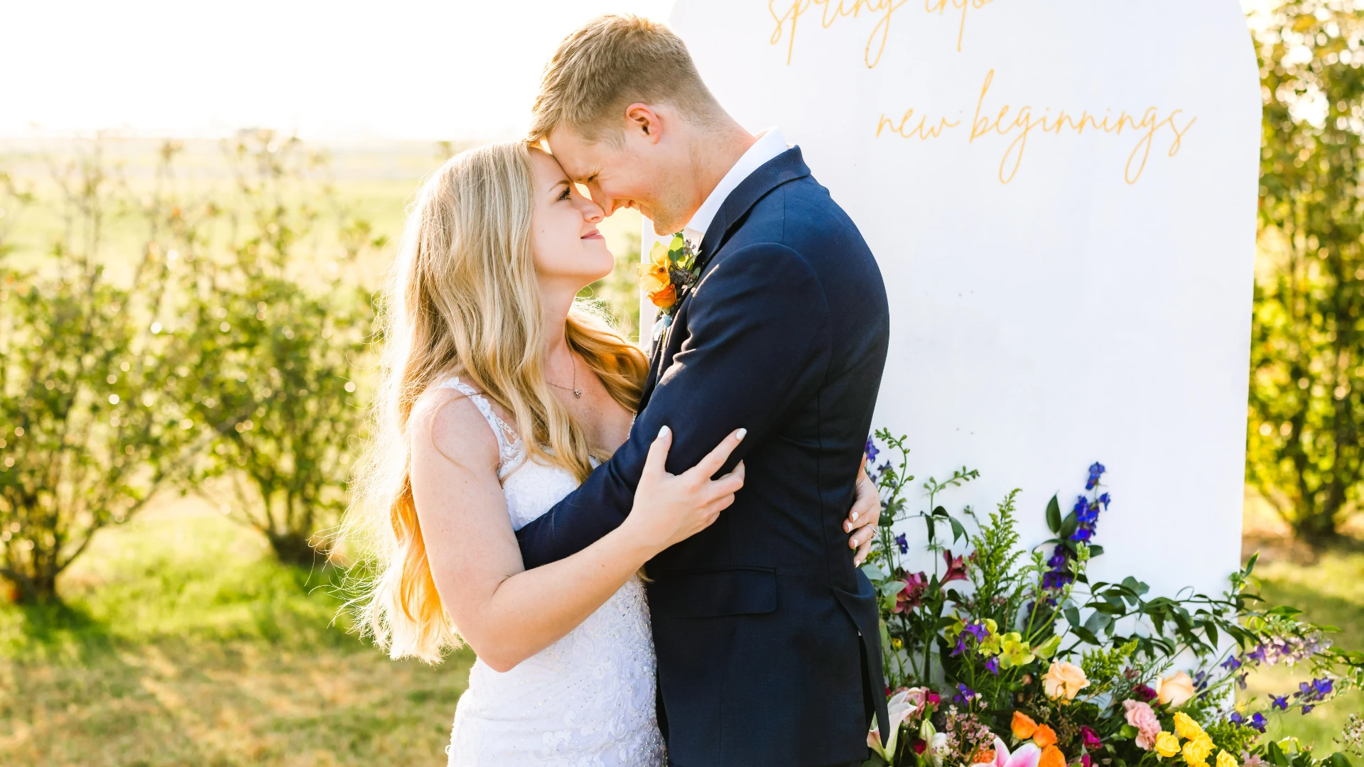 A bride and groom embrace and touch foreheads, standing in front of an outdoor floral arrangement.