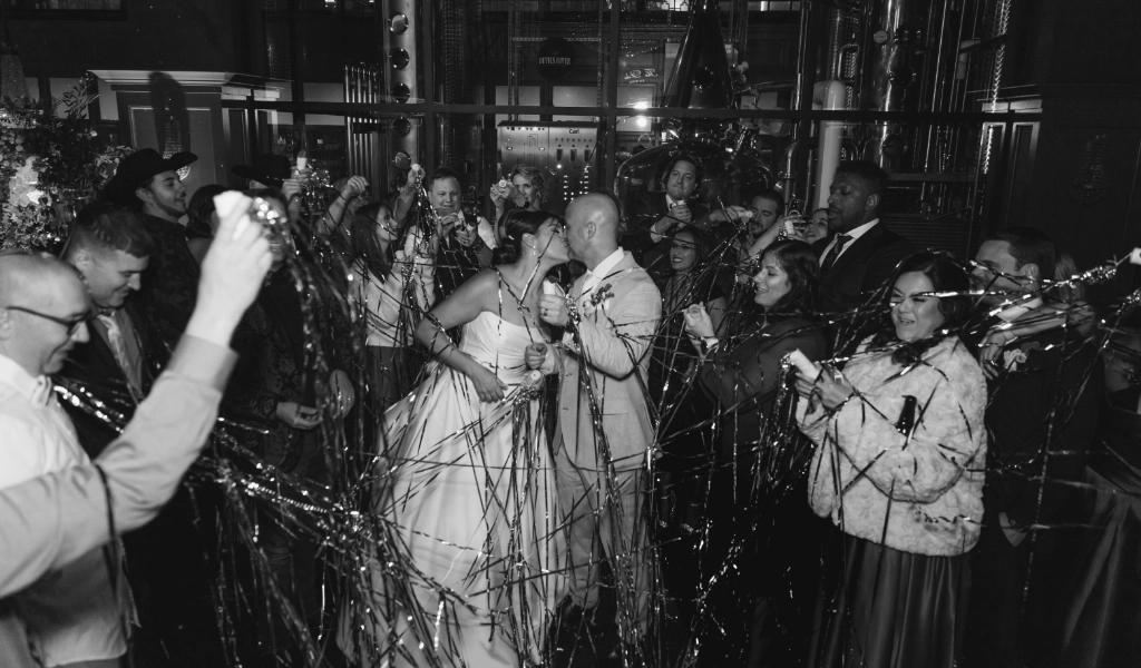A bride and groom kiss while surrounded by a crowd of people waving streamers in a festive celebration in a dimly lit venue, perfectly orchestrated by expert wedding planning services.