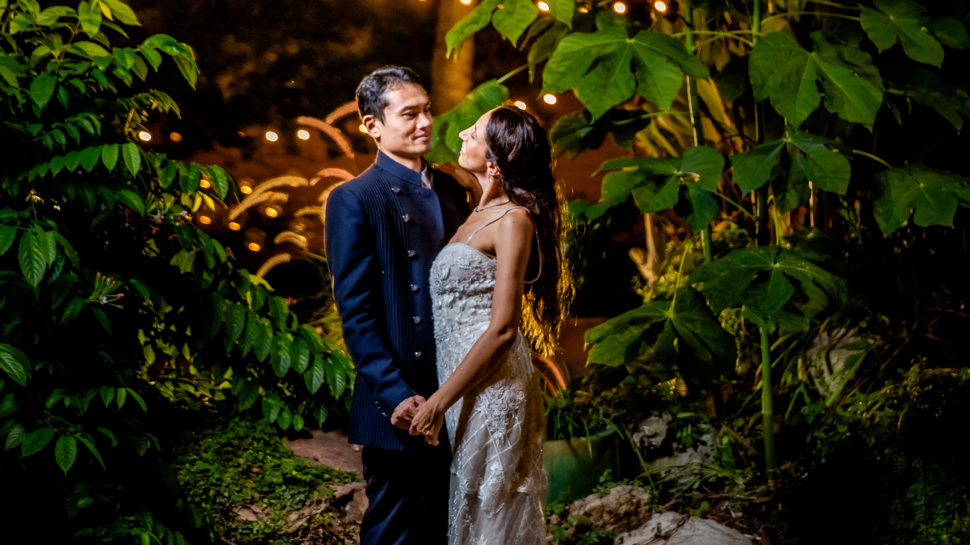 A couple dressed in formal attire stand closely together, gazing at each other amidst lush greenery and soft, warm lighting, as their wedding planner ensures every detail is perfect.
